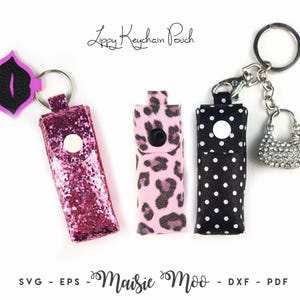 May include: Three rectangular keychain pouches with snap closures. One is pink glitter with a lip-shaped charm, one is pink and black leopard print, and one is black with white polka dots and a rhinestone handbag charm. Text reads Lippy Keychain Pouch.