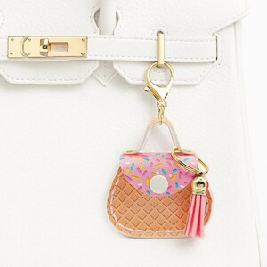 Peut inclure: Un porte-cl&eacute;s en forme de mini-sac &agrave; main avec un motif de paillettes roses et blanches sur le rabat, un corps textur&eacute; en gaufre marron clair et un pompon rose. Il est attach&eacute; &agrave; un sac &agrave; main en cuir blanc avec des accessoires dor&eacute;s.