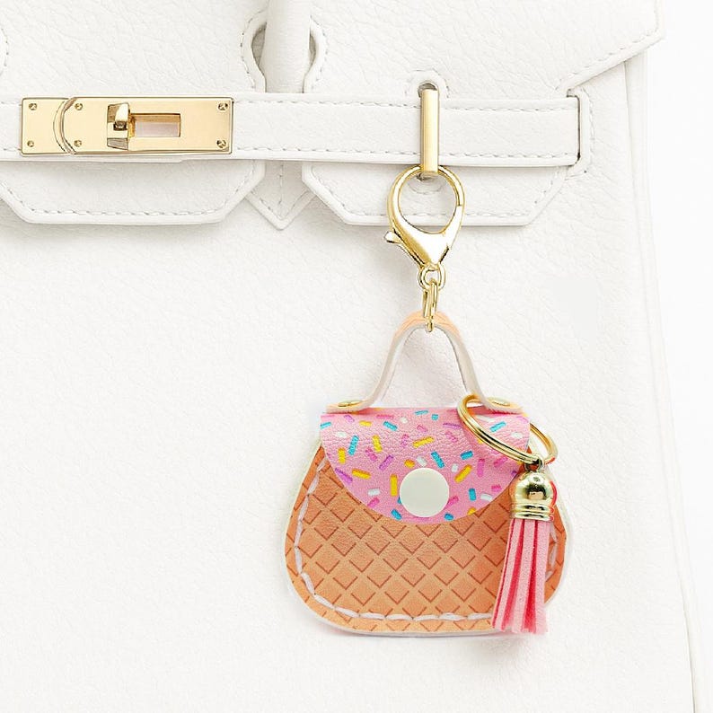 Peut inclure: Un porte-cl&eacute;s en forme de mini-sac &agrave; main avec un motif de paillettes roses et blanches sur le rabat, un corps textur&eacute; en gaufre marron clair et un pompon rose. Il est attach&eacute; &agrave; un sac &agrave; main en cuir blanc avec des accessoires dor&eacute;s.