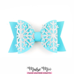 May include: A bright turquoise faux leather hair bow featuring two large, silver glittery snowflake cutouts layered on each side. The bow has a textured surface and a central band, with the text Maisie Moo Faux Leather Crafts below it.