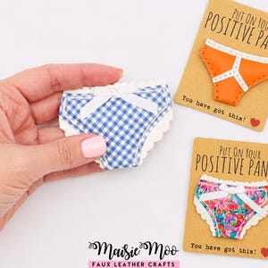 May include: Three faux leather panty-shaped crafts. One blue and white gingham pattern, one solid orange, and one floral print. Each features a white bow and scalloped edges. Two are on cards reading "PUT ON YOUR POSITIVE PANTS You have got this!"