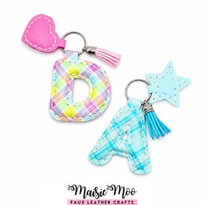 Puede incluir: Dos llaveros con forma de letra en piel sintética cosida. Uno es una letra D a cuadros con un corazón rosa y una borla rosa claro. El otro es una letra A a cuadros con una estrella azul claro y una borla azul brillante. El texto dice Maisie Moo.