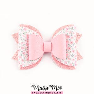 May include: A layered pink faux leather bow with a floral print and glitter accents. The bow features a central knot and is designed with multiple layers. The text "Maisie Moo Faux Leather Crafts" is at the bottom.