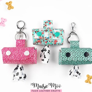 Op de afbeelding: Drie rechthoekige poepzakjeshouders van kunstleer met metalen sleutelhangers. Eén is roze glitter met witte drukknopen, één heeft een bloemenprint met honden en turquoise drukknopen, en één is turquoise met zwarte stippen en zwarte drukknopen.