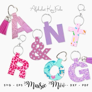 May include: A collection of alphabet key fobs featuring letters A, N, R, O, G, an ampersand, and a cross shape. The keychains display various patterns including pink floral, purple texture, confetti, pink leopard print, polka dots, and purple glitter.