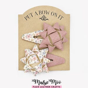 May include: Two hair clips on a brown card labelled PUT A BOW ON IT and Maisie Moo Faux Leather Crafts. One clip features a dusty pink bow with a floral patterned base, while the other has a floral patterned bow with a solid dusty pink base.