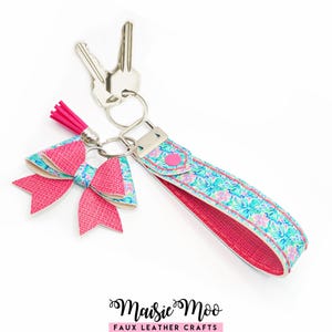 Può includere: Un portachiavi con un cinturino da polso e un fiocco decorativo, entrambi realizzati in materiale con motivo floreale nei toni del turchese, rosa e bianco. Il cinturino ha un interno rosa acceso e il set include una nappa rosa e due chiavi argentate.
