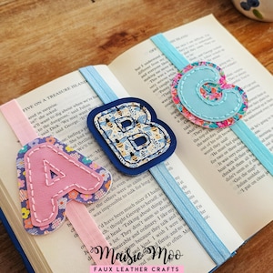 Puede incluir: Tres marcadores de tela con las letras A, B y C. La letra A es rosa con un estampado floral, la B tiene un estampado de abejas y la C es azul claro. Cada marcador está unido a una cinta, descansando sobre las páginas de un libro abierto.