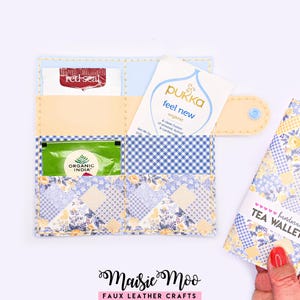 May include: A rectangular tea wallet made of faux leather with a blue and yellow floral and gingham patchwork pattern. The wallet features multiple interior pockets holding tea bags from Red Seal, Pukka, and Organic India, secured by a snap closure.