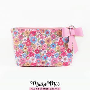 Puede incluir: Un estuche rectangular con cremallera que presenta un vibrante estampado floral con flores rosas, azules, naranjas y blancas sobre un fondo claro. Un lazo rosa claro liso está unido al tirador de la cremallera. El texto Maisie Moo Faux Leather Crafts aparece debajo.