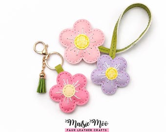Op de afbeelding: Drie bloemvormige sleutelhangers van kunstleer in de kleuren roze, lichtroze en lavendel. Elke bloem heeft een geel hart met witte stiksels en een groen bandje. Een goudkleurige karabijnhaak en een klein groen kwastje zijn bevestigd.