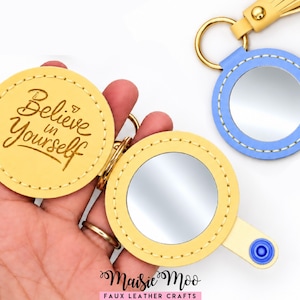 May include: A yellow faux leather circular keychain mirror with the text Believe in Yourself embossed on the cover. A second blue circular mirror keychain with a gold ring and tassel is visible. Both items feature white stitching.