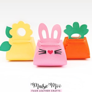 May include: Three colourful faux leather Easter-themed gift boxes. A yellow flower-shaped box, a pink bunny-faced box, and an orange carrot-shaped box. Each box has a unique cutout design against a white background.