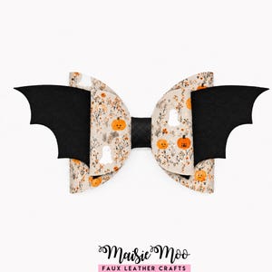 May include: A hair bow featuring a beige faux leather material with a pattern of orange pumpkins, white ghosts, and brown foliage. The bow includes two black bat-shaped wings extending from the sides and a solid black center knot. The text Maisie Moo FAUX LEATHER CRAFTS is printed at the bottom.