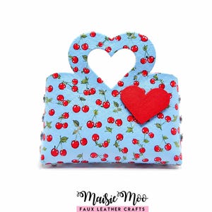 May include: A light blue handbag featuring a repeating cherry pattern and a heart-shaped cutout handle. A solid red heart applique is attached to the front. The text Maisie Moo FAUX LEATHER CRAFTS is printed in black below the handbag.