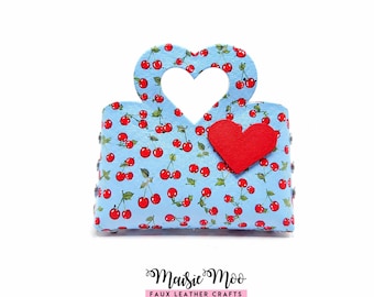 Op de afbeelding: Een lichtblauwe handtas met een herhalend kersenpatroon en een hartvormige uitgesneden handgreep. Een effen rood hart is op de voorkant bevestigd. De tekst Maisie Moo FAUX LEATHER CRAFTS staat in het zwart onder de handtas gedrukt.