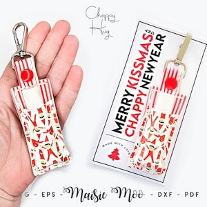 May include: A lip balm holder featuring a red and white striped pattern with small elf figures. It includes a silver metal swivel clasp and a red snap button. The attached display card reads MERRY KISSMAS AND CHAPPY NEWYEAR.