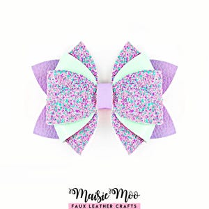 Peut inclure: Un nœud pour cheveux multicouche composé de similicuir lavande texturé, de panneaux vert menthe lisses et de sections pailletées dans des tons de violet, rose et bleu sarcelle. Le centre est fixé par une bande lavande. Texte : Maisie Moo.