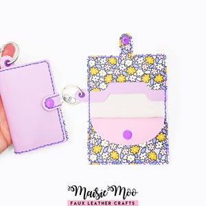 Può includere: Un portacarte in similpelle viola chiaro con chiusura a scatto e anello in metallo. Accanto, un portacarte aperto mostra un motivo floreale di fiori gialli e bianchi su sfondo azzurro, dotato di linguetta a scatto e anello metallico.