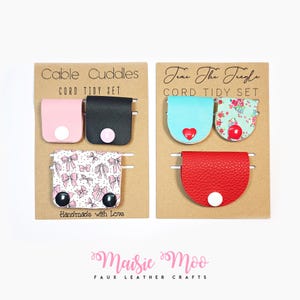 Peut inclure: Ensembles de range-cordons Cable Cuddles et Time The Jingle. Chaque ensemble comprend plusieurs organiseurs de câbles en similicuir de différentes couleurs et motifs, dont rose, noir, bleu, floral et rouge. Le texte "Handmade with Love" est visible.