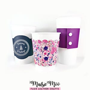 以下が含まれることがあります： 取り外し可能なスリーブが付いた3つの白いカップ。左のスリーブはダークブルーで、白い円形の「Season's Greetings」のグラフィックが描かれています。中央のスリーブはスカラップエッジで、ピンクの花柄と2つのダークボタンが付いています。右のスリーブはパープルです。