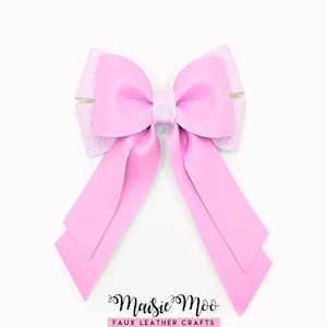 Pode incluir: Um laço decorativo com um centro de couro sintético rosa claro liso e duas pontas longas e afiadas. O laço está sobreposto a uma base mais larga de cor rosa claro brilhante. O texto Maisie Moo FAUX LEATHER CRAFTS está impresso em preto abaixo.
