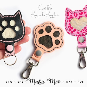 May include: Three cat fur keepsake keychains. One black cat-shaped keychain with white paw prints, one peach paw print keychain, and one pink leopard print cat-shaped keychain with a heart cutout. Each keychain has a metal clasp.