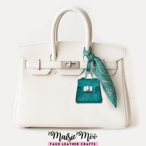 May include: A white faux leather handbag with silver hardware, a teal purse charm, and a teal and gold paisley scarf. The handbag has a top handle and a rectangular shape. The charm and scarf add a pop of color to the neutral bag.
