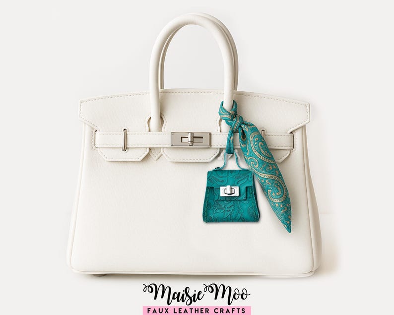 May include: A white faux leather handbag with silver hardware, a teal purse charm, and a teal and gold paisley scarf. The handbag has a top handle and a rectangular shape. The charm and scarf add a pop of color to the neutral bag.