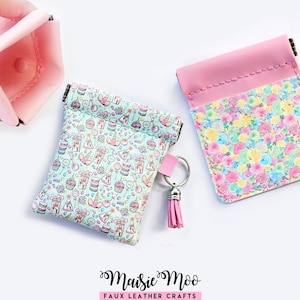 May include: A collection of small, colorful pouches. One is light blue with a pattern of sweets and unicorns, another is pink with a floral design. A pink pouch is held by a hand. The pouches are made of faux leather.