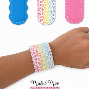 May include: A wristband featuring a rainbow-colored animal print pattern on faux leather with white stitching. Above it are three additional faux leather strips in solid blue, rainbow animal print, and solid pink, all with scalloped edges.