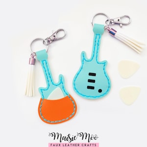 May include: Two guitar-shaped keychains made of faux leather with turquoise and orange accents, featuring white tassels, silver-toned lobster clasps, and two white guitar picks. The text Maisie Moo Faux Leather Crafts is visible at the bottom.