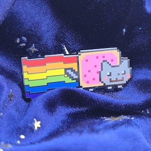 Puede incluir: Un colorido pin de esmalte con un diseño pixelado de Nyan Cat. El pin muestra un gato gris con un cuerpo de Pop-Tart rosa, un rastro de arcoíris y una bandera arcoíris. El pin está sobre un fondo azul oscuro.