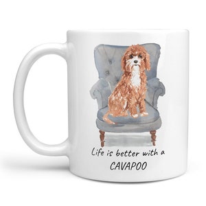 Könnte beinhalten: Weiße Keramiktasse mit einer Aquarell-Illustration eines braunen und weißen Cavapoo-Hundes, der in einem blauen Sessel sitzt. Der Text "Life is better with a CAVAPOO" ist unter dem Bild gedruckt.
