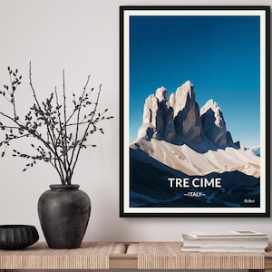 Könnte beinhalten: Ein gerahmter Kunstdruck mit einer Berglandschaft unter einem klaren blauen Himmel. Der Druck trägt die Aufschrift "TRE CIME - ITALY" in weißem Text. Das Kunstwerk wird auf einem Holztisch mit dekorativen Gegenständen ausgestellt.