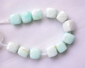 May include: A strand of light blue and white gemstone beads, each bead is faceted and has a square shape. The beads are strung on a white cord.