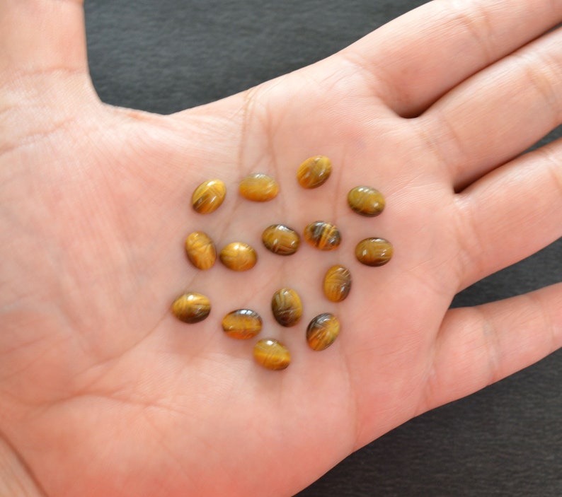 Puede incluir: Una colecci&oacute;n de trece peque&ntilde;as piedras preciosas de ojo de tigre de forma ovalada. Cada piedra presenta una superficie lisa y pulida con bandas chatoyantes caracter&iacute;sticas de color marr&oacute;n dorado y oscuro que crean un efecto brillante.