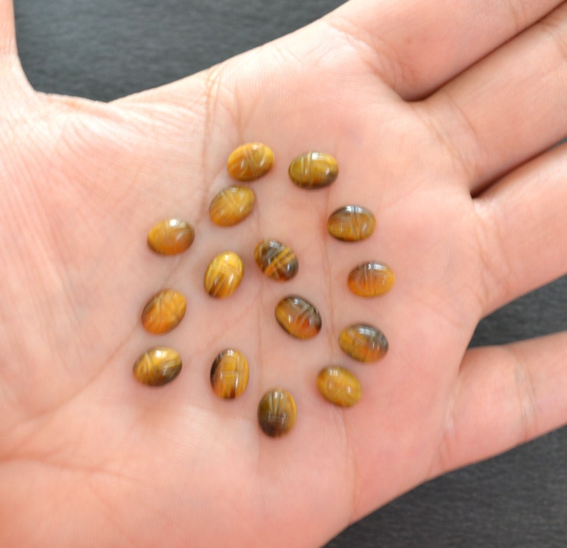 Puede incluir: Una colecci&oacute;n de trece peque&ntilde;as piedras preciosas de ojo de tigre de forma ovalada, cada una con un dise&ntilde;o de escarabajo tallado en la superficie. Estas piedras pulidas de color marr&oacute;n y amarillo dorado muestran un brillo natural.