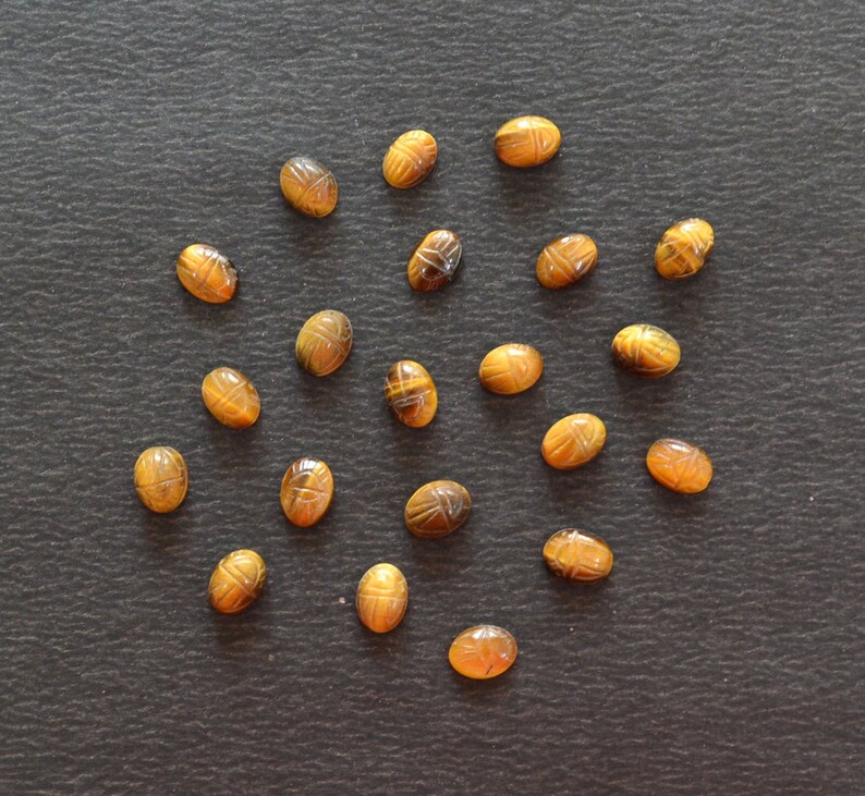 Puede incluir: Una colecci&oacute;n de diecinueve piedras preciosas de ojo de tigre de forma ovalada, cada una tallada con un dise&ntilde;o detallado de escarabajo. Estas piedras pulidas de color marr&oacute;n y amarillo dorado presentan bandas naturales.