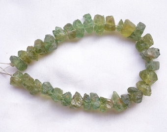 May include: A strand of raw, unpolished green gemstone chips arranged in a teardrop shape. The stones vary in size and shape, featuring translucent to opaque textures with natural facets and inclusions, strung together on a thin silver-colored wire.