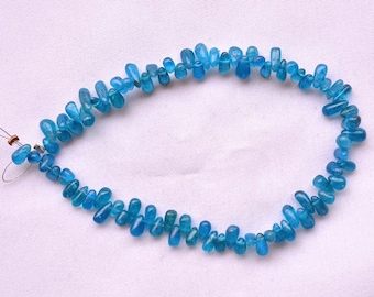 Puede incluir: Un hilo de pequeñas cuentas de apatita azul en forma de lágrima dispuestas en un patrón circular. Las piedras preciosas translúcidas de color azul vibrante están ensartadas en un alambre fino, con los extremos del alambre visibles a la izquierda.