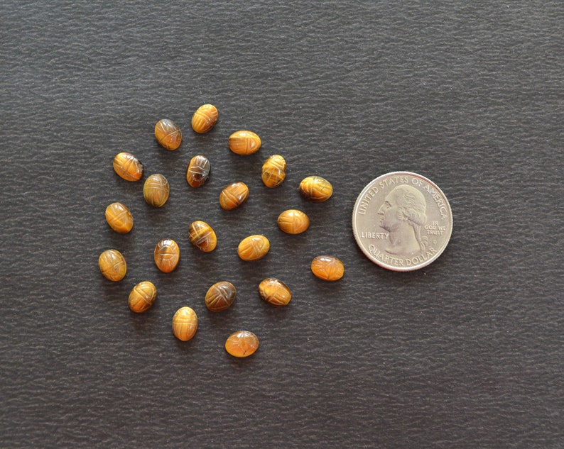 Puede incluir: Una colecci&oacute;n de diecinueve peque&ntilde;as piedras preciosas de ojo de tigre de forma ovalada talladas con un dise&ntilde;o de escarabajo, dispuestas junto a una moneda de veinticinco centavos de EE. UU. para mostrar la escala y el tama&ntilde;o.