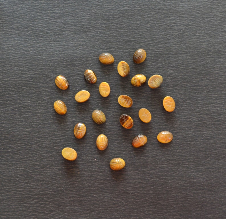 Puede incluir: Una colecci&oacute;n de diecinueve peque&ntilde;as piedras preciosas de ojo de tigre de forma ovalada, cada una con un dise&ntilde;o de escarabajo tallado en la superficie. Las piedras muestran bandas naturales de color marr&oacute;n dorado y chatoyance, dispuestas en un grupo.
