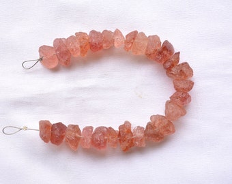 May include: A strand of rough-cut, peach-colored gemstones, strung on a thin wire. The stones vary in size and shape, with a slightly translucent appearance. The background is a plain white fabric.