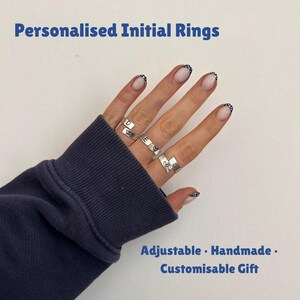 Peut inclure: Bagues réglables argentées avec les initiales D, E et M, chacune ornée d'un cœur. Les bagues sont présentées sur une main, avec le texte "Personalised Initial Rings" en haut et "Adjustable . Handmade . Customisable Gift" en bas.