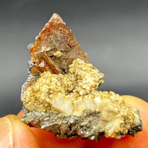 May include: A close-up of a mineral specimen featuring a prominent, orange-brown crystal formation atop a matrix of smaller, granular, light-yellow crystals. The base is a mix of gray and silver-coloured minerals. The overall structure is approximately 5 cm in length.