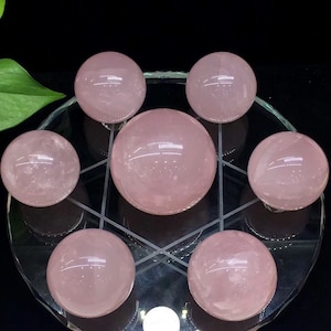 May include: Seven pink rose quartz spheres arranged in a geometric pattern on a clear glass surface. The spheres are smooth and polished, with a light pink hue.