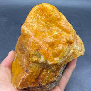 May include: A rough, uncut stone with a vibrant orange and yellow hue. The surface has a textured, uneven appearance, with areas of lighter and darker coloration. The stone is held in a hand, showcasing its natural form and size.