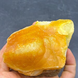 Könnte beinhalten: Ein roher, ungeschliffener Edelstein mit einem leuchtenden gelb-orangenen Farbton. Die Oberfläche hat ein strukturiertes Aussehen mit Farbintensitätsvariationen. Der Stein wird in einer Hand gehalten und zeigt seine natürliche, unregelmäßige Form. Ideal für Sammler.