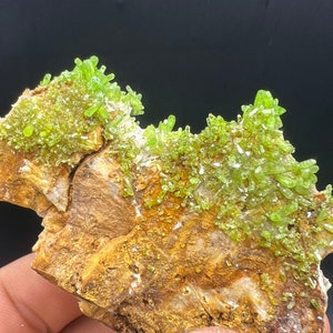 Op de afbeelding: Een cluster van groene kristallen op een bruine rots. De kristallen zijn scherp en puntig en zijn in een willekeurig patroon gerangschikt. De rots is ruw en getextureerd.